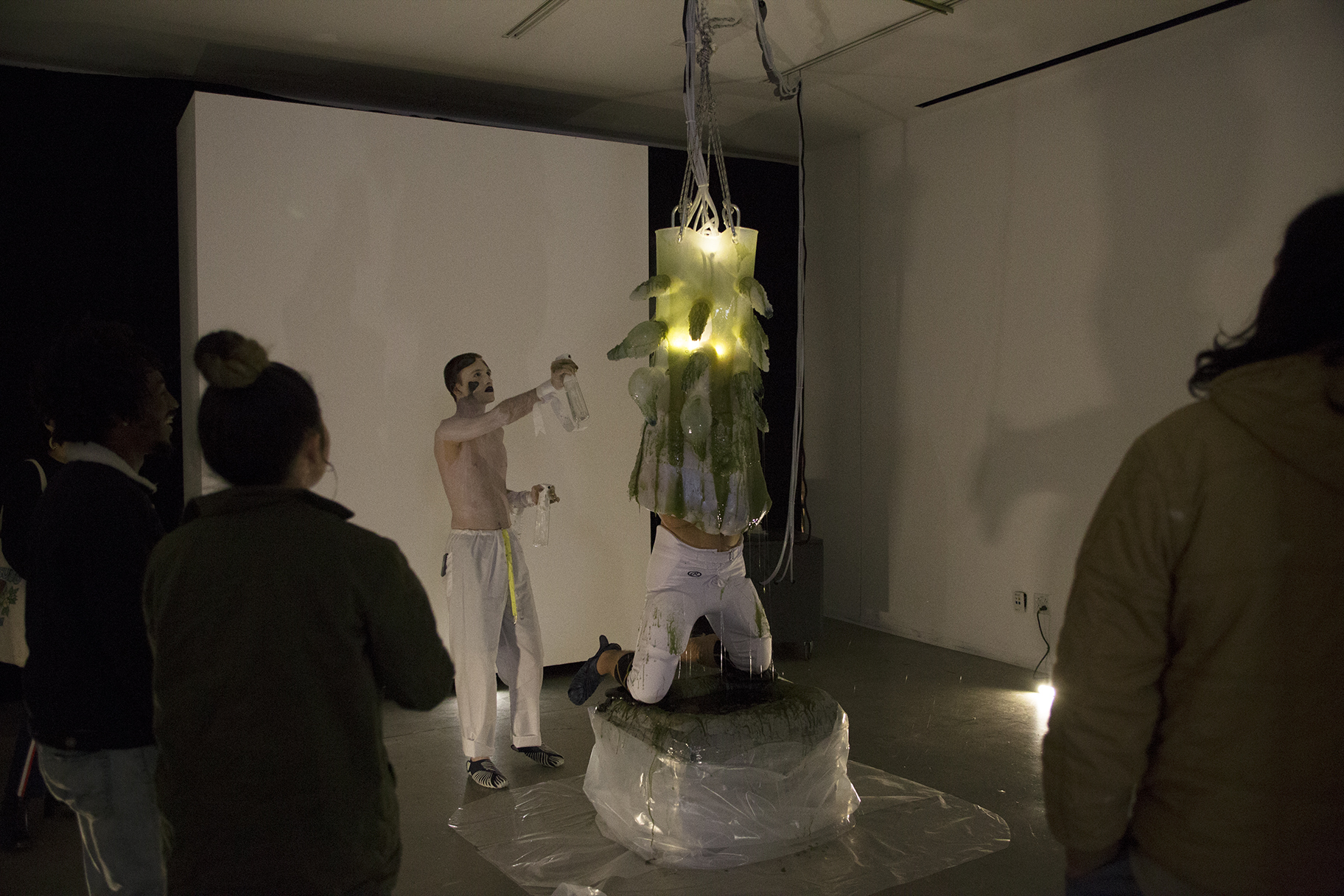 A man engulfed in a gelatenous form covered in tentacles knealing over a pool of green slime while a shirtless man painted in white sprays him with moisture while a crowd watches